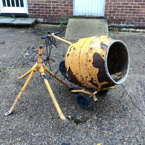 Vintage cement mixer with tripod stand in a garden setting.