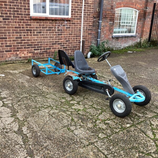 Blue pedal go kart with trailer in a garden setting.