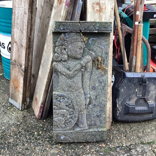 Antique stone wall plaque with carved religious figure in garden setting.