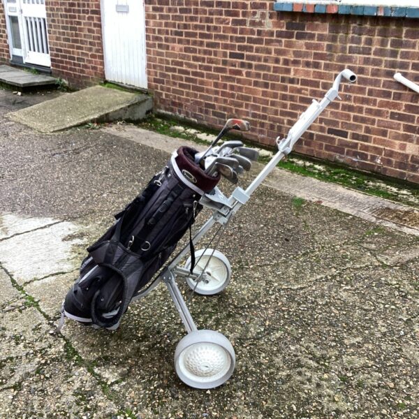 Black golf bag with silver folding trolley on pavement.