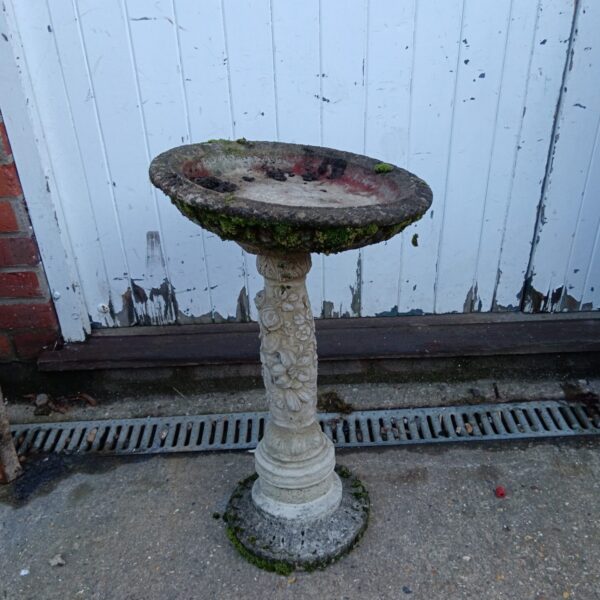 Weathered concrete bird bath on a fluted plinth in a garden.
