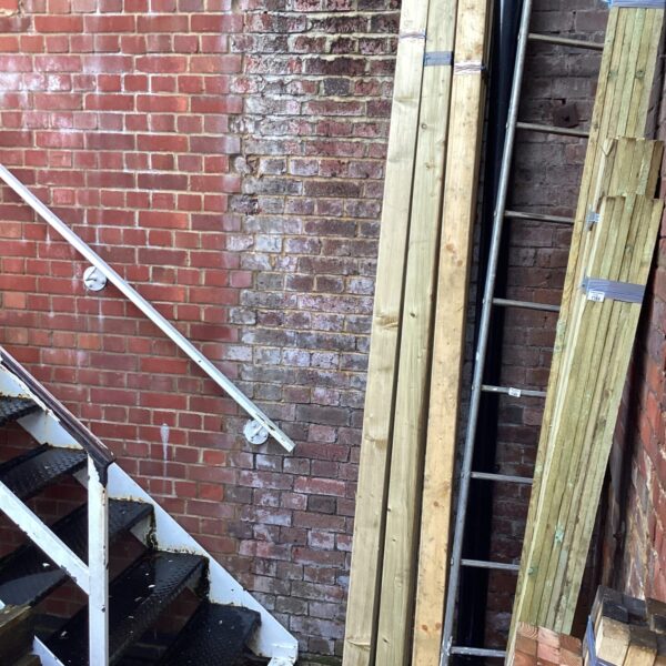 Bundles of 9ft timber leaning against a brick wall in a garden.