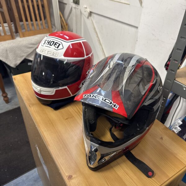 Pair of Takachi and Shoei motorsport helmets on a wooden surface.