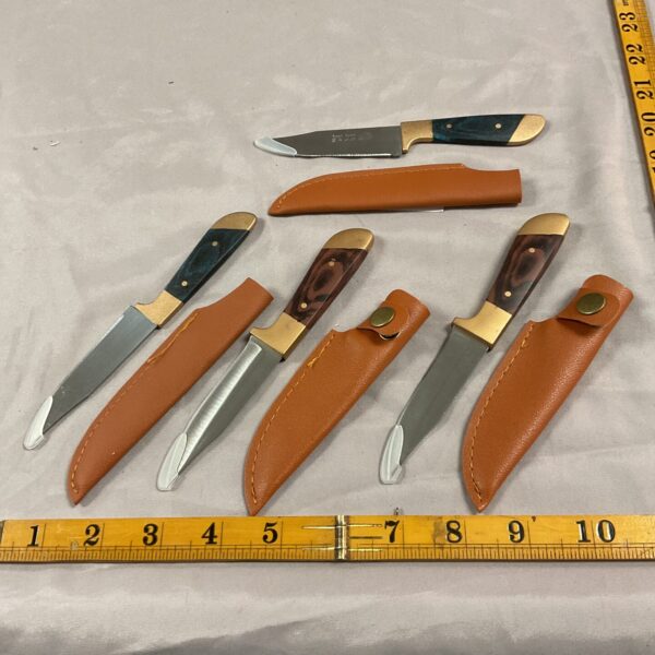 Four vintage sheath knives with leather sheaths displayed on a ruler.