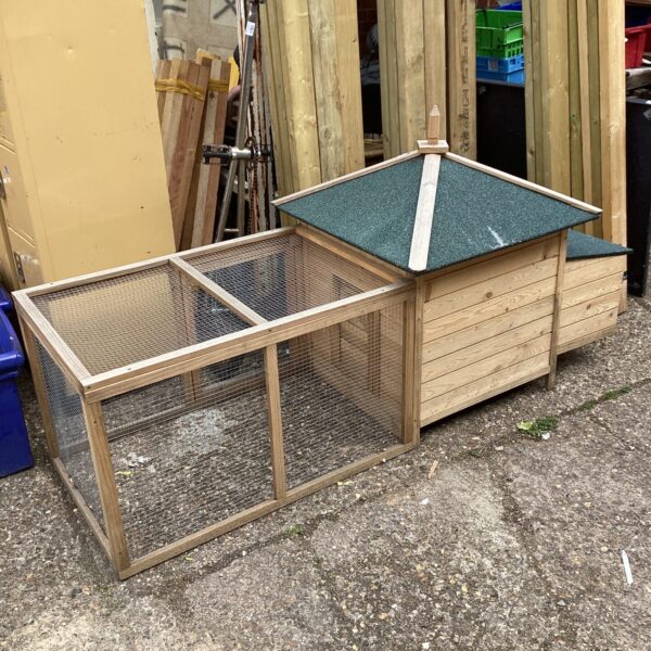 Large wooden chicken coop with wire mesh panels in a garden setting.