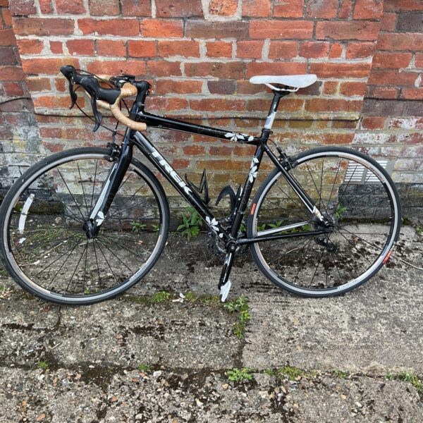 Side view of a black Trek Lexa S road bike against a brick wall.