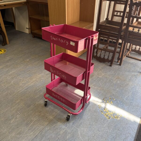 Pink metal storage cart with three tiers and wheels.
