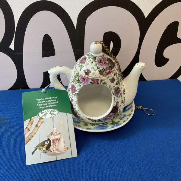 Delicate porcelain teapot bird feeder with floral design in a garden setting.