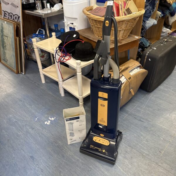 Sebo Automatic X4 vacuum cleaner with spare bags shown in a cluttered room.